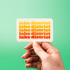 Hand holding a custom vinyl sticker with 'Lake District' text on a green background.
