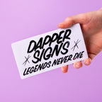 Hand holding a custom vinyl sticker with 'Dapper Signs Legends Never Die' text against a purple background.