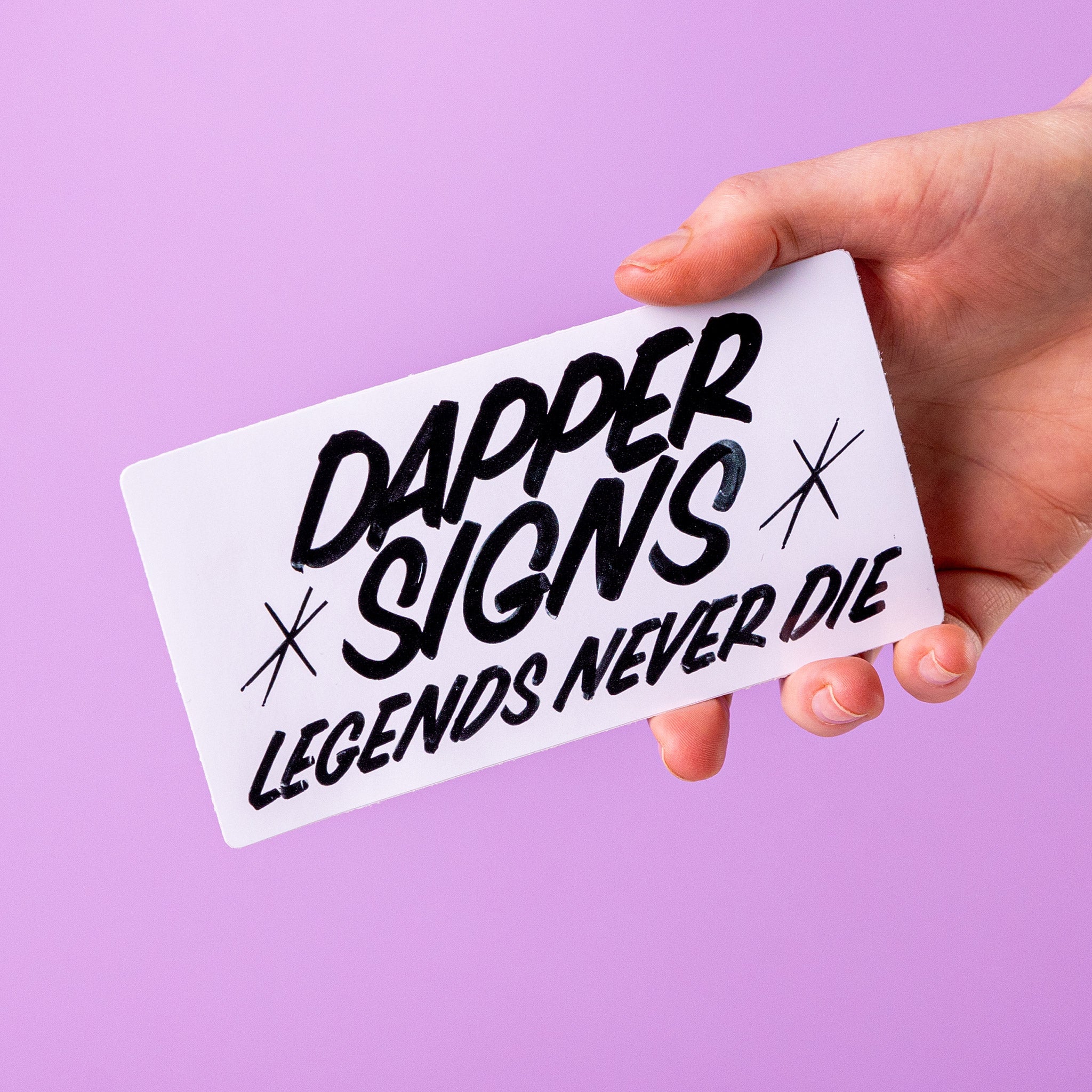 Hand holding a custom vinyl sticker with 'Dapper Signs Legends Never Die' text against a purple background.