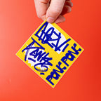 Hand holding a yellow and blue custom vinyl sticker with text against an orange background.