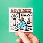 Custom vinyl sticker with 'LOVESICK TATTOO' design held by a hand against a green background.